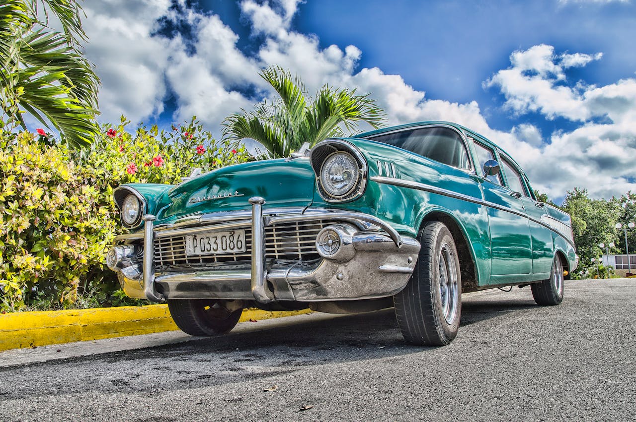 A beautifully restored classic car on a sunny road in Havana, Cuba, with lush foliage and a vibrant sky.